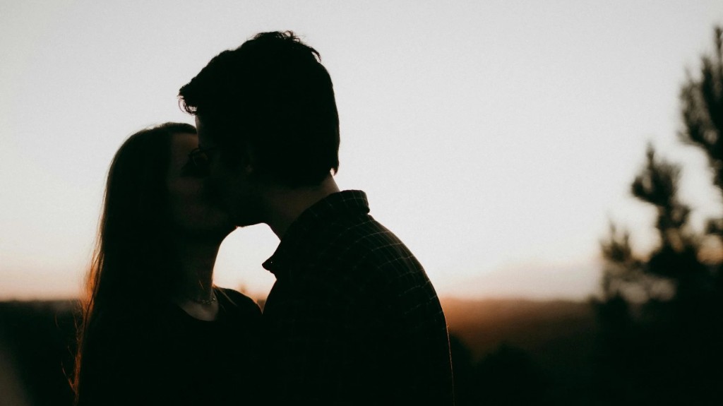 Couple in silhouette sharing a kiss against a bright, warm sunset sky.